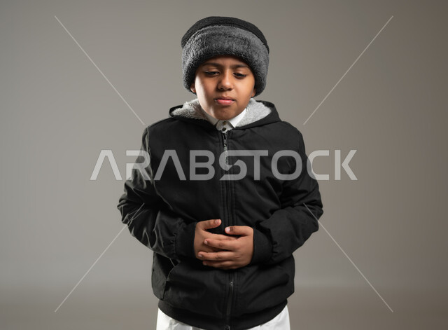 وضع اليدين على البطن و النظر للكاميرا بإيماءات تدل على الألم والامتعاض, بورتريه مقرب لولد عربي خليجي سعودي يرتدي الثوب التقليدي وملابس الشتاء الدافئة يحتاج إلى العلاج والراحة, خلفية رمادية, محتوى حساس, استخدام حساس