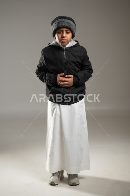 الشعور بالتعب والإرهاق والمغص , بورتريه لولد عربي سعودي خليجي يرتدي الثوب التقليدي وملابس الشتاء الدافئة بايماءات وجه ويدين تدل على الشعور بألم في البطن الحاجه للعلاج و الدواء, خلفية رمادية، الجسم بالطول الكامل، استخدام حساس، محتوى حساس