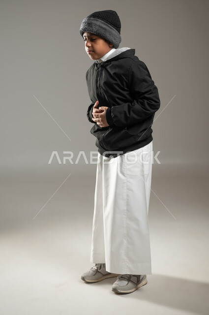 الشعور بالمغص و التعب والإرهاق, بورتريه لولد عربي سعودي خليجي يرتدي الثوب التقليدي وملابس الشتاء الدافئة بايماءات وجه ويدين تدل على الشعور بألم في البطن الحاجه للعلاج و الدواء, خلفية رمادية، الجسم بالطول الكامل، استخدام حساس، محتوى حساس