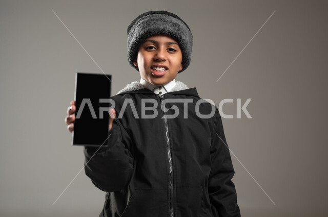 استخدام التطبيقات الحديثة و التسويق الألكتروني, بورتريه مقرب لولد عربي سعودي خليجي مبتسم  يرتدي الثوب التقليدي وملابس الشتاء الدافئة يمسك بهاتف محمول بشاشة سوداء فارغة, برامج التواصل الاجتماعي عبر الهاتف المحمول, خلفية رمادية، محتوى حساس، استخدام حساس