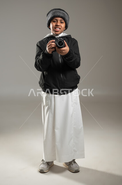استخدام التقنيات الحديثة, التقاط صور تذكارية عبر الكاميرا,  بورتريه لولد عربي خليجي سعودي يرتدي الثوب التقليدي وملابس الشتاء الدافئة ينظر بايماءات السرور, موهبة واحتراف فن التصوير, صورة الجسم بالطول الكامل, خلفية رمادية، محتوى حساس، استخدام حساس