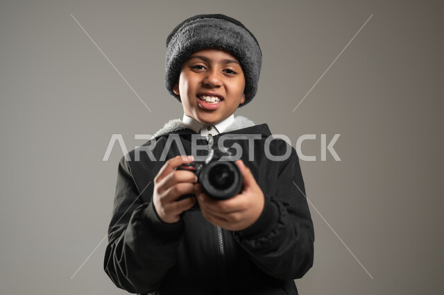 استخدام  الاجهزة الحديثة, التقاط صور تذكارية عبر الكاميرا,  بورتريه مقرب لولد عربي خليجي سعودي يرتدي الثوب التقليدي وملابس الشتاء الدافئة ينظر بايماءات السرور, موهبة واحتراف فن التصوير, خلفية رمادية، محتوى حساس، استخدام حساس