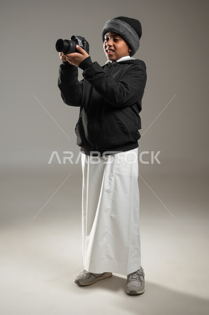 التقاط صور تذكارية عبر الكاميرا, استخدام التقنيات الحديثة, بورتريه لولد عربي خليجي سعودي يرتدي الثوب التقليدي وملابس الشتاء الدافئة يقوم بتصوير شيء ما, صور عالية الجودة, موهبة واحتراف فن التصوير, صورة الجسم بالطول الكامل, خلفية رمادية، محتوى حساس، استخدام حساس
