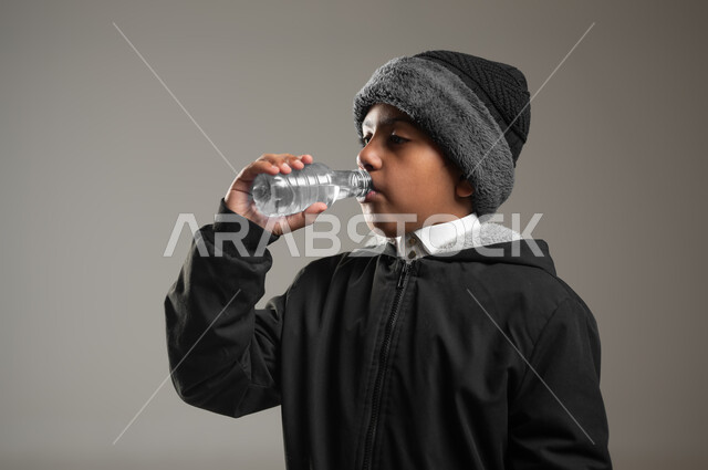 التخلص من سموم الجسم، أهمية شرب الماء النقي للمحافظة على بشرة نضرة، بورتريه مقرب لولد عربي خليجي سعودي يرتدي الثوب التقليدي و ملابس الشتاء الدافئة، يقوب بشرب المياه من عبوة بلاستيكية، المحافظة على سلامة الجهاز المناعي، خلفية رمادية، استخدام حساس، محتوى حساس