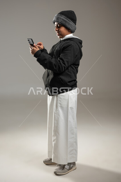 Requesting communication via mobile phone, social networking via mobile phone, portrait of a Saudi Arabian Gulf boy wearing a traditional dress and warm winter clothes, facial and hand gestures indicating a personal conversation, full length body, gray background, sensitive content, sensitive use