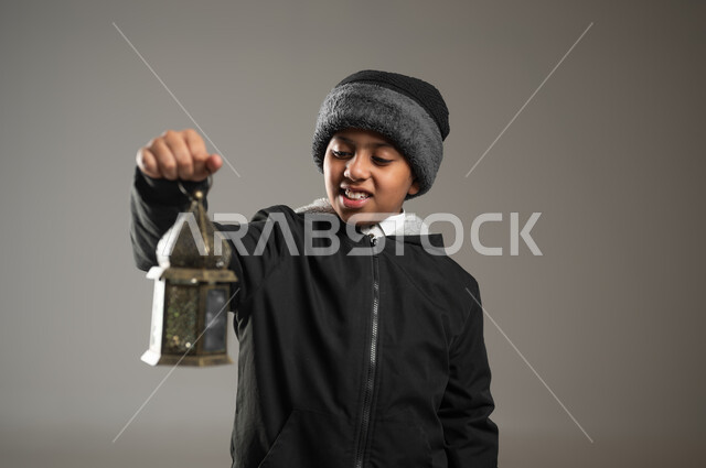 الإمساك بالفانوس الجميل المزخرف والنظر إليه بإيماءات البهجة, بورتريه مقرب  لطفل عربي خليجي سعودي يرتدي الثوب التقليدي، التحضير لدخول شهر رمضان, زينة الأعياد والمناسبات الإسلامية, خلفية رمادية، محتوى حساس, استخدام حساس