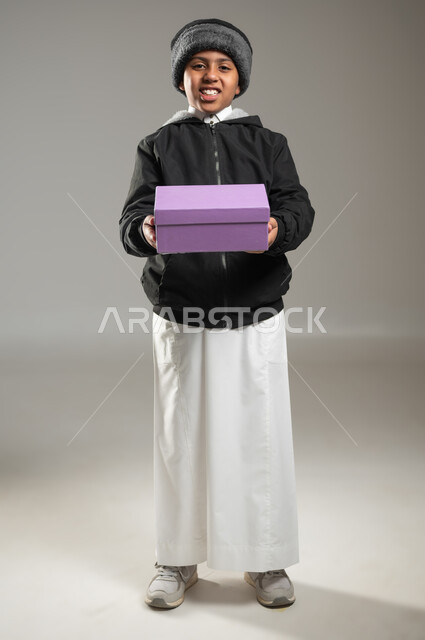 Gestures and facial and hand expressions indicating happiness, readiness to give gifts, portrait of a Saudi Gulf Arab child wearing a traditional dress and winter clothing, standing and holding a colorful gift box in his hands, looking at the camera with gestures of pleasure, full-length body, gray background, sensitive content, sensitive use
