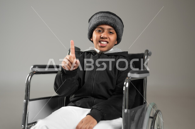 اهتمام ودعم المجتمع السعودي لذوي الاحتياجات الخاصه, بورتريه مقرب لطفل عربي سعودي خليجي يرتدي الثوب التقليدي ولباس الشتاء يجلس على كرسي متحرك يشير بإصبعه للاعلى , النظر الى الكاميرا بإيماءات السرور، خلفية رمادية، محتوى حساس، استخدام حساس