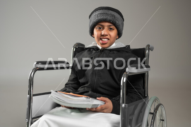 The Saudi community’s interest and support for people with special needs, a portrait of a Saudi Gulf Arab child wearing the traditional dress and winter clothing, sitting in a wheelchair holding his school books in his hand, looking at the camera with gestures of pleasure, gray background, sensitive content, sensitive use