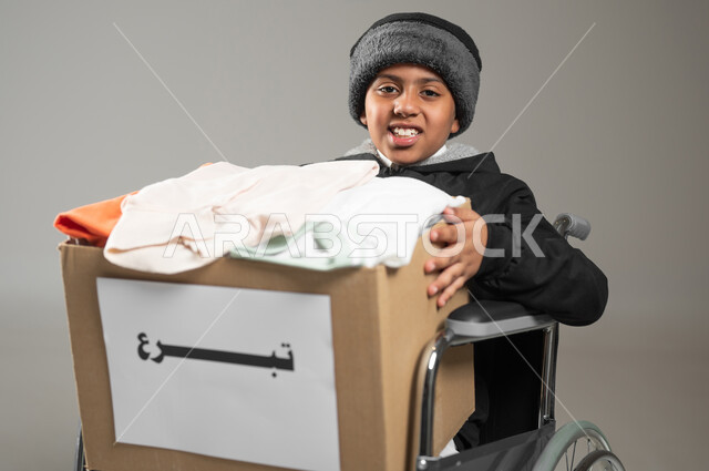التبرع للجمعيات السعودية، التصدق بالفائض من المستلزمات، بورتريه مقرب لولد عربي خليجي سعودي يرتدي الثوب التقليدي ولباس الشتاء يجلس على كرسي متحرك يحمل بيديه صندوق كرتوني مليء بالملابس ينظر الى الكاميرا بايماءات السرور، خلفية رمادية, محتوى حساس, استخدام حساس