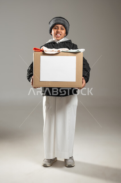 جمع التبرعات للفقراء والمساكين, جمعيات خيرية و انسانية، بورتريه لطفل عربي خليجي سعودي يرتدي الثوب التقليدي ولباس الشتاء يحمل صندوق من الملابس ينظر الى الكاميرا بايماءات الحاجه للمال والمساعدة ، الجسم بالطول الكامل، خلفية رمادية, محتوى حساس, استخدام حساس