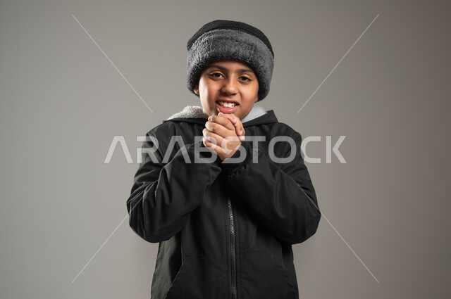 The extreme cold of winter, a close-up portrait of a Gulf Arab boy wearing the traditional dress and winter clothes, holding his palms together due to the extreme cold, looking at the camera with extreme cold gestures, clothes suitable for the winter, winter weather in Saudi Arabia, gray background, sensitive content, sensitive use