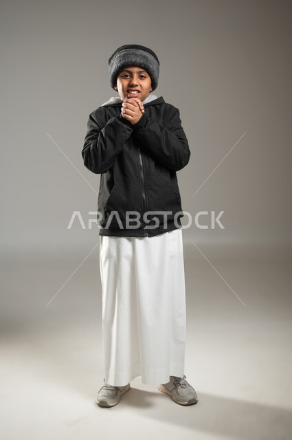 النظر الى الكاميرا بإيماءات البرد الشديد، بورتريه  لولد عربي خليجي يرتدي الثوب التقليدي و ملابس الشتاء يضم كفيه من شدة البرودة،  ملابس مناسبة لفصل الشتاء، الأجواء الشتوية في السعودية، البرد القارس لفصل الشتاء، الجسم بالطول الكامل، خلفية رمادية, محتوى حساس , استخدام حساس