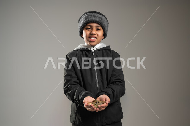 المساعدات والتبرعات الإنسانية، التبرع للجمعيات السعودية، بورتريه مقرب لطفل عربي خليجي سعودي يرتدي الثوب التقليدي و ملابس الشتاء يحمل بيده كومة من العملات المعدنية، الأعمال الخيرية الإسلاميه، خلفية رمادية, محتوى حساس , استخدام حساس