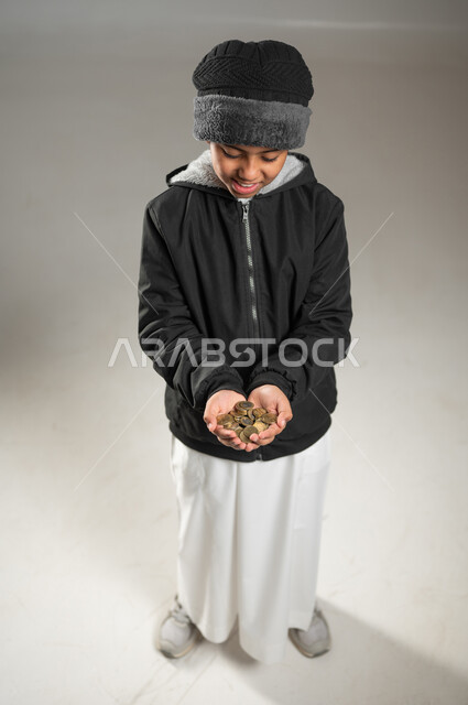 التبرع للجمعيات السعودية، ايماءات الفقر والحاجه للمال والمساعدة، بورتريه لطفل عربي خليجي سعودي يرتدي الثوب التقليدي و ملابس الشتاء يحمل بيده كومة من العملات المعدنية، الجسم بالطول الكامل، خلفية رمادية , محتوى حساس , استخدام حساس