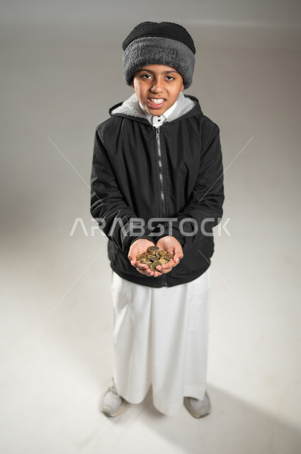 النظر الى الكاميرا بتعابير الحزن والحاجة، التبرع للجمعيات السعودية، بورتريه لطفل عربي خليجي سعودي يرتدي الثوب التقليدي و ملابس الشتاء يحمل بيده كومة من العملات المعدنية، الجسم بالطول الكامل، خلفية رمادية , محتوى حساس , استخدام حساس