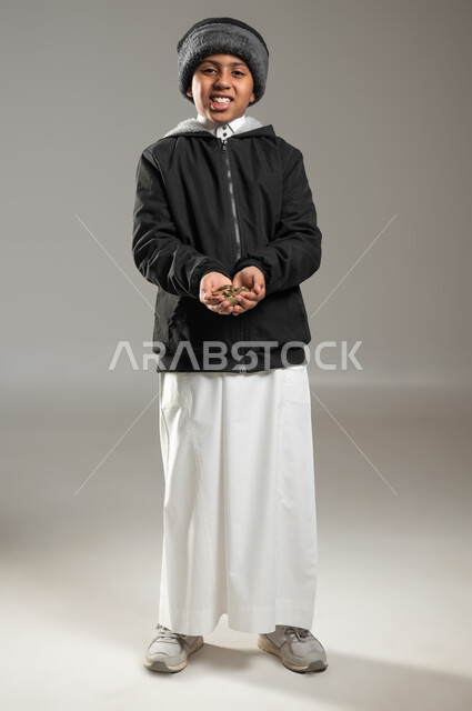 مفهوم الاحتياج وعدم امتلاك الاموال، ايماءات الفقر والحاجه للمال والمساعدة، بورتريه لطفل عربي خليجي سعودي يرتدي الثوب التقليدي يحمل بيده كومة من العملات المعدنية، تعابير الحزن والحاجة، الجسم بالطول الكامل، خلفية رمادية , محتوى حساس , استخدام حساس