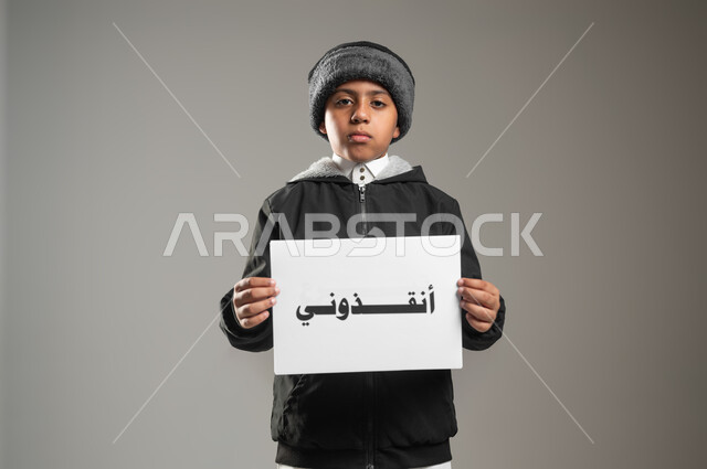 المساعدات والتبرعات الإنسانية، الأعمال الخيرية الإسلاميه، بورتريه لطفل عربي سعودي خليجي يرتدي الثوب التقليدي وملابس الشتاء يمسك بيده ورقه مكتوب بها انقذوني، النظر إلى الكاميرا بايماءات الفقر والحاجه للمال والمساعدة، خلفية رمادية، استخدام حساس، محتوى حساس
