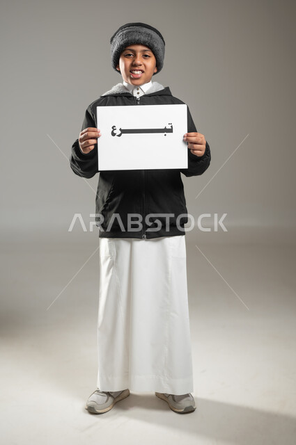 جمعيات خيرية اسلاميه، مبادرات إنسانية وتطوعية ، مفهوم الأعمال الخيرية الإسلاميه والتبرع  في السعودية، بورتريه لطفل عربي سعودي خليجي يرتدي الثوب التقليدي وملابس الشتاء يمسك بيده ورقه مكتوب بها تبرع، الجسم بالطول الكامل، خلفية رمادية، استخدام حساس، محتوى حساس