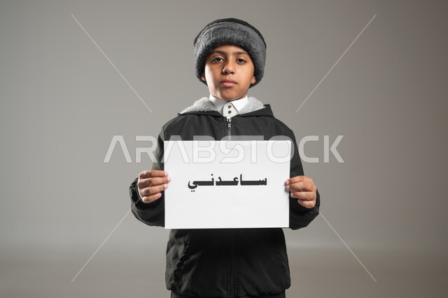 مساعدة المحتاجين والفقراء، مبادرات إنسانية وتطوعية خيرية، مفهوم الأعمال الخيرية الإسلاميه والتبرع  في السعودية، بورتريه مقرب لطفل عربي سعودي خليجي يرتدي الثوب التقليدي وملابس الشتاء يمسك بيده ورقه مكتوب بها ساعدني، خلفية رمادية، استخدام حساس، محتوى حساس