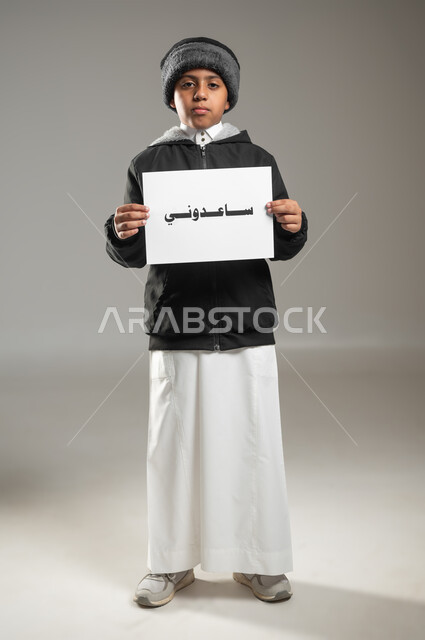 Islamic charitable works, helping the poor, poor and needy, humanitarian and charitable volunteer initiatives, portrait of a Saudi Gulf Arab child wearing traditional dress and winter clothing holding in his hand a paper with the words Help Me written on it, full length body, gray background, sensitive use, sensitive content