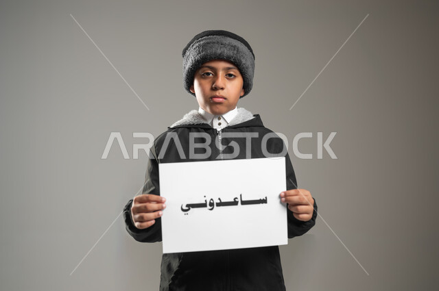 مبادرات إنسانية وتطوعية خيرية،  مساعدة الفقراء والمحتاجين، مفهوم الأعمال الخيرية الإسلاميه والتبرع  في السعودية، بورتريه مقرب لطفل عربي سعودي خليجي يرتدي الثوب التقليدي وملابس الشتاء يمسك بيده ورقه مكتوب بها ساعدوني، خلفية رمادية، استخدام حساس، محتوى حساس