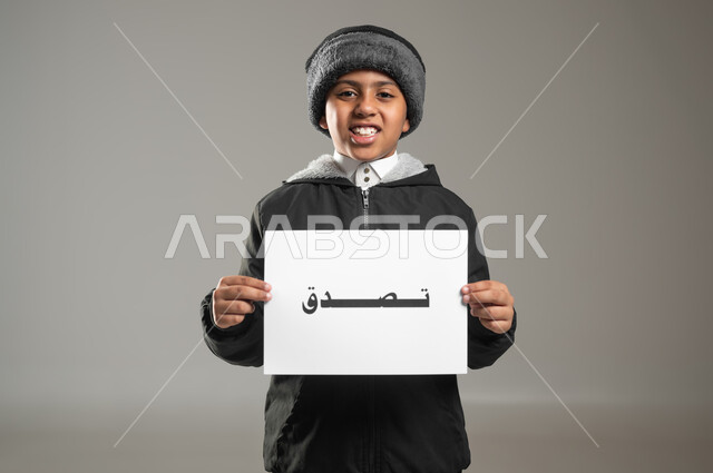 مساعدة المساكين والفقراء والمحتاجين، مفهوم الأعمال الخيرية الإسلاميه والتبرع  في السعودية، بورتريه مقرب لطفل عربي سعودي خليجي يرتدي الثوب التقليدي وملابس الشتاء يمسك بيده ورقه مكتوب بها تصدق، خلفية رمادية، استخدام حساس، محتوى حساس