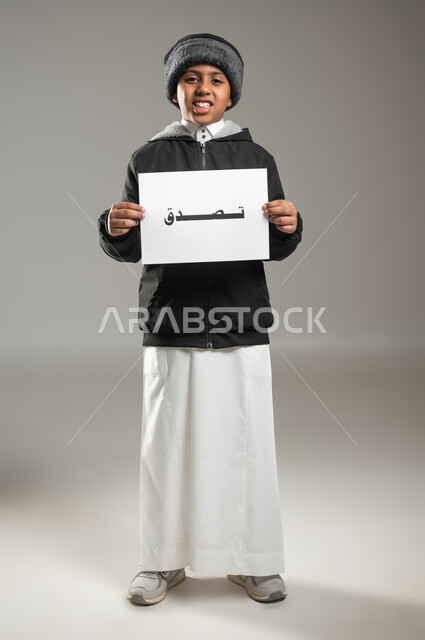 مساعدة المساكين والفقراء والمحتاجين، مبادرات إنسانية وتطوعية خيرية، بورتريه لطفل عربي سعودي خليجي يرتدي الثوب التقليدي وملابس الشتاء يمسك بيده ورقه مكتوب عليها تصدق، الأعمال الخيرية الإسلاميه، الجسم بالطول الكامل، خلفية رمادية، استخدام حساس، محتوى حساس