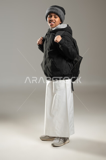 النظر إلى الكاميرا بإيماءات السرور والسعادة، عودة العام الدراسي الجديد في المملكة، بورتريه لطالب عربي خليجي سعودي مبتسم يرتدي الثوب التقليدي وملابس الشتاء الدافئة يحمل حقيبه الظهر المدرسيه، الجسم بالطول الكامل، خلفيه رمادية، محتوى حساس، استخدام حساس