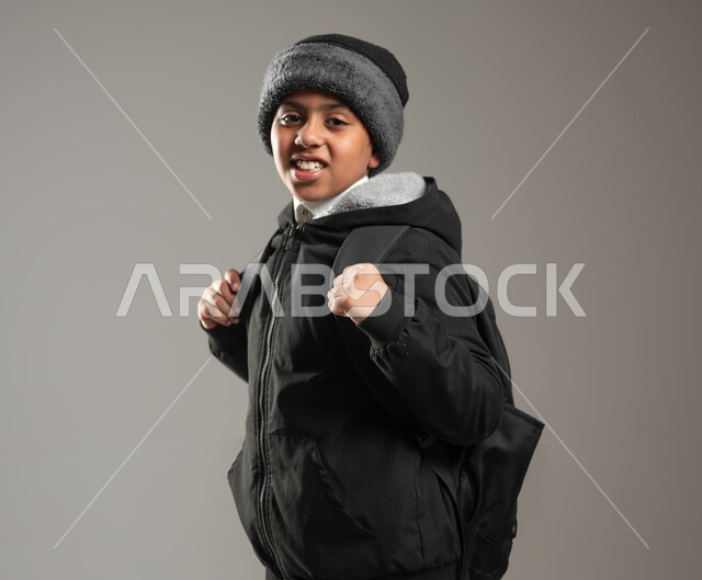 الوقوف باستقامة والنظر إلى الكاميرا بإيماءات السرور, بداية عام دراسي جديد، بورتريه مقرب لطالب عربي خليجي سعودي يرتدي الثوب التقليدي وملابس الشتاء الدافئة يحمل حقيبه الظهر المدرسيه، استقبال العام الدراسي الجديد بحماس، خلفيه رمادية، محتوى حساس، استخدام حساس