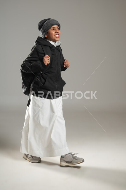 Gestures of pleasure for returning to school, preparing to go to school, beginning of a new school year, portrait of a smiling Saudi Arabian Gulf student wearing the traditional dress and warm winter clothes holding a school backpack, full-length body, gray background, sensitive content, sensitive use