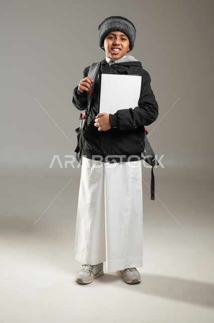 الاستعداد للذهاب الى المدرسة، بداية عام دراسي جديد، بورتريه لطالب عربي خليجي سعودي مبتسم يرتدي الثوب التقليدي وملابس الشتاء الدافئة يحمل حقيبه الظهر المدرسيه، ايماءات السرور للعودة الى الدراسة، الجسم بالطول الكامل، خلفيه رمادية، محتوى حساس، استخدام حساس