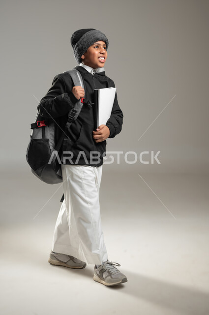 بداية عام دراسي جديد، الاستعداد للذهاب الى المدرسة، بورتريه لطالب عربي خليجي سعودي مبتسم يرتدي الثوب التقليدي وملابس الشتاء الدافئة يحمل حقيبه الظهر المدرسيه، ايماءات السرور للعودة الى الدراسة، الجسم بالطول الكامل، خلفيه رمادية، محتوى حساس، استخدام حساس