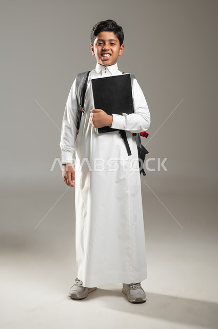 النظر الى الكاميرا بإيماءات السرور والفرح, تعابير البشاشة والاستعداد للذهاب الى المدرسة, صورة لولد عربي خليجي سعودي يرتدي حقيبة الظهر ويحمل بيده مجموعة من الملفات, مفهوم التعليم الدراسي في السعودية، الجسم بالطول الكامل، خليفة رمادية، محتوى حساس، استخدام حساس