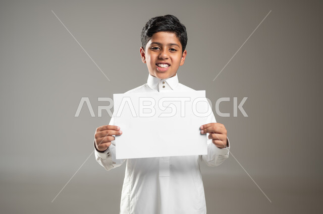 Collecting and distributing charitable donations, helping the poor, the poor and the needy, humanitarian aid and donations, a close-up portrait of a Saudi Gulf Arab child wearing a traditional dress and holding a blank piece of paper in his hand, Islamic charitable work in Saudi Arabia, gray background, sensitive use, sensitive content