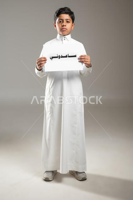 مساعدة المساكين والفقراء والمحتاجين، مفهوم التبرع والأعمال الخيرية الإسلاميه في السعودية، بورتريه لطفل عربي سعودي خليجي يرتدي الثوب التقليدي و يمسك بيده ورقه مكتوب عليها ساعدوني، الجسم بالطول الكامل، خلفية رمادية، استخدام حساس، محتوى حساس