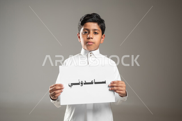 مبادرات وتوزيع الاحتياجات للمحتاجين، النظر إلى الكاميرا بايماءات الفقر والحاجه للمال والمساعدة، بورتريه مقرب لطفل عربي سعودي خليجي يرتدي الثوب التقليدي و يمسك بيده ورقه مكتوب عليها ساعدوني، الأعمال الخيرية الإسلاميه، خلفية رمادية، استخدام حساس، محتوى حساس