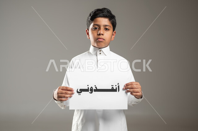 المساعدات والتبرعات الإنسانية، الأعمال الخيرية الإسلاميه، بورتريه مقرب لطفل عربي سعودي خليجي يرتدي الثوب التقليدي يمسك بيده ورقه مكتوب بها انقذوني، النظر إلى الكاميرا بايماءات الفقر والحاجه للمال والمساعدة، خلفية رمادية، استخدام حساس، محتوى حساس