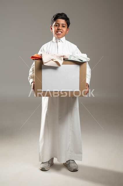 جمع وتوزيع التبرعات للفقراء والمساكين, جمعيات خيرية ومبادرات انسانية،  بورتريه لطفل عربي خليجي سعودي يرتدي الثوب التقليدي يحمل صندوق من الملابس ينظر الى الكاميرا بايماءات الفقر والحاجه للمال والمساعدة ، الجسم بالطول الكامل، خلفية رمادية, محتوى حساس, استخدام حساس