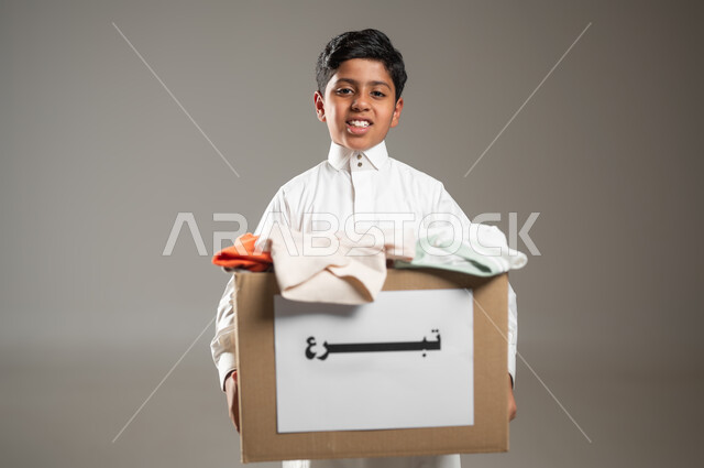 جمعيات خيرية ومبادرات انسانية، جمع وتوزيع التبرعات للفقراء والمساكين, بورتريه مقرب لطفل عربي خليجي سعودي يرتدي الثوب التقليدي يحمل صندوق من الملابس ينظر الى الكاميرا بايماءات الفقر والحاجه للمال والمساعدة ، خلفية رمادية, محتوى حساس, استخدام حساس