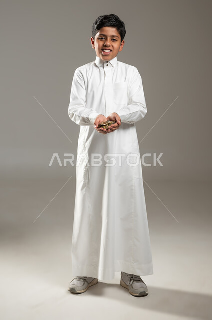 مفهوم الاحتياج وعدم امتلاك الاموال، تعابير الحزن والحاجة، بورتريه لطفل عربي خليجي سعودي يرتدي الثوب التقليدي يحمل بيده كومة من العملات المعدنية، ايماءات الفقر والحاجه للمال والمساعدة ، الجسم بالطول الكامل، خلفية رمادية , محتوى حساس , استخدام حساس