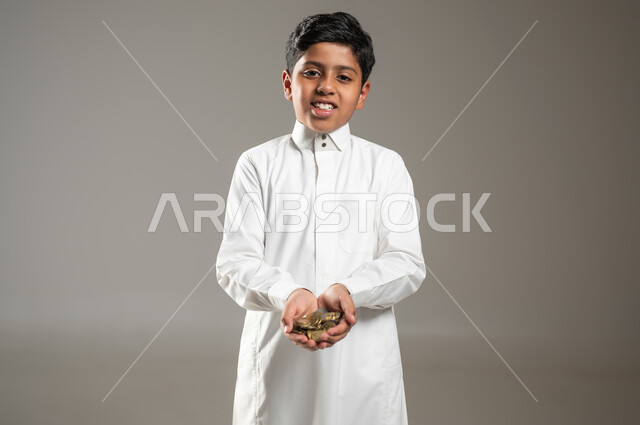 تعابير الحزن والحاجة، مفهوم الاحتياج وعدم امتلاك الاموال، بورتريه مقرب لطفل عربي خليجي سعودي يرتدي الثوب التقليدي يحمل بيده كومة من العملات المعدنية، ايماءات الفقر والحاجه للمال والمساعدة ، خلفية رمادية , محتوى حساس , استخدام حساس