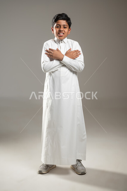 التعبير عن المشاعر والاحتياجات بحركات لغة الجسد, ضم اليدين نحو الكتفين بشكل متقاطع دلالة على الحب والاحتواء, النظر إلى الكاميرا بتعبيرات الابتسام والسعادة, بورتريه لفتى عربي خليجي سعودي يرتدي الثوب التقليدي, إشارات ودلالات لها معنى, جسم بالطول الكامل, خلفية رمادية, محتوى حساس, استخدام حساس