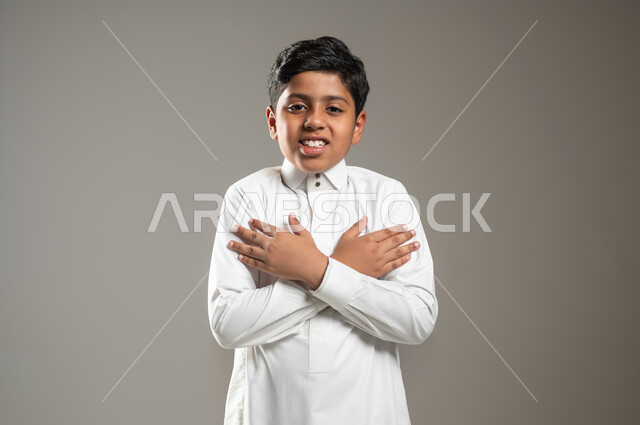 ضم اليدين نحو الكتفين بشكل متقاطع, النظر إلى الكاميرا بتعبيرات الابتسام والسعادة, بورتريه لفتى عربي خليجي سعودي يرتدي الثوب التقليدي, حركات لغة الجسد, إشارات ودلالات يتم التعبير عنها, خلفية رمادية, محتوى حساس, استخدام حساس
