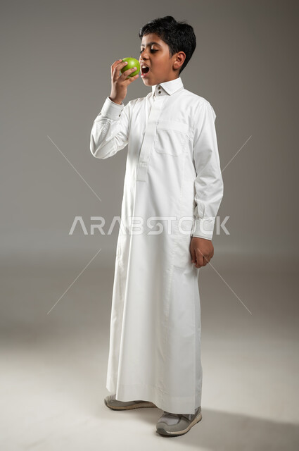 فاكهة لذيذة وشهية, طعم التفاح اللذيذ, دور الطعام الصحي في بناء الجسم, بورتريه لولد عربي خليجي سعودي يرتدي الزي الأبيض التقليدي يحمل بيده  تفاحة خضراء, الاهتمام بالصحة, جسم بالطول الكامل, خلفية رمادية, محتوى حساس, استخدام حساس