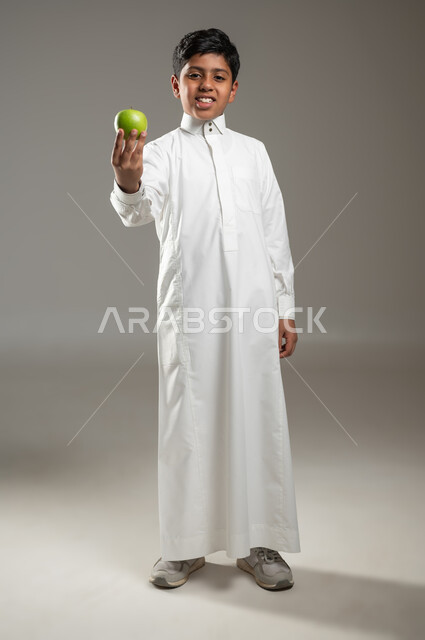 The importance of the nutrients found in apples, a healthy mind in a healthy body, portrait of a Saudi Gulf Arab boy wearing traditional white clothing holding a delicious apple in his hand and looking at the camera with happy gestures, full-length body, gray background, sensitive content, sensitive use
