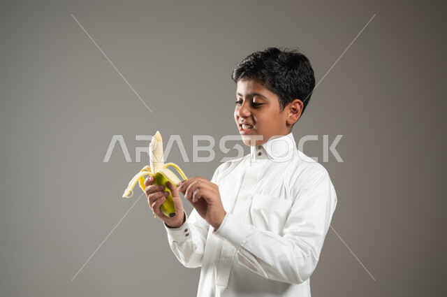 إتباع نظام غذائي صحي متوازن, وجبة فطور صحية طازجة, بورتريه مقرب لولد عربي خليجي سعودي مبتسم يرتدي الثوب التقليدي يمسك بالموزة ويقوم بتقشيرها, مفهوم العناية والاهتمام بالصحة, خلفية رمادية, محتوى حساس, استخدام حساس