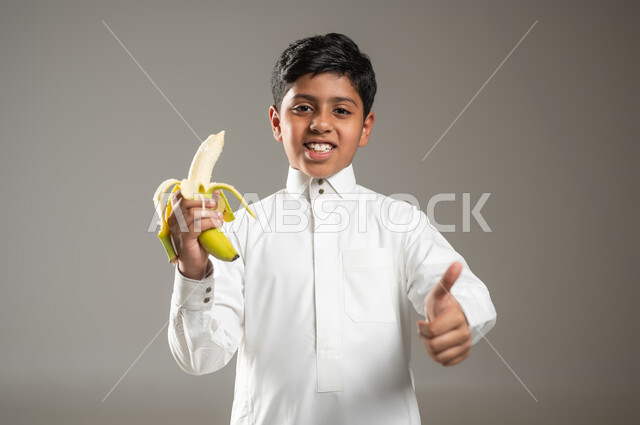 النظر إلى الكاميرا ورفع الاصبع باشارة تدل على السعادة, إتباع نظام غذائي صحي, بورتريه مقرب لولد عربي خليجي سعودي مبتسم يرتدي الزي الأبيض التقليدي يحمل بيده موزة, مفهوم العناية والاهتمام بالصحة, خلفية رمادية, محتوى حساس, استخدام حساس