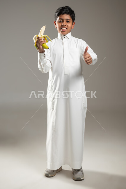 وجبة فطور صحية طازجة, إتباع نظام غذائي صحي, بورتريه لولد عربي خليجي سعودي مبتسم يرتدي الزي الأبيض التقليدي يحمل بيده موزة, النظر إلى الكاميرا و الإشارة بالاصبع بإيماءات السرور والسعادة, جسم بالطول الكامل, خلفية رمادية, محتوى حساس, استخدام حساس