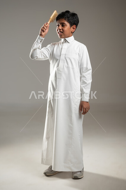الاهتمام بالمظهر الخارجي, بورتريه لولد عربي  خليجي سعودي يرتدي الثوب السعودي التقليدي ويستخدم فرشاة الشعر, تصفيف الشعر، تعابير وجه تدل على السعادة، العناية بالجمال, جسم بالطول الكامل، خلفية رمادية, محتوى حساس، استخدام حساس